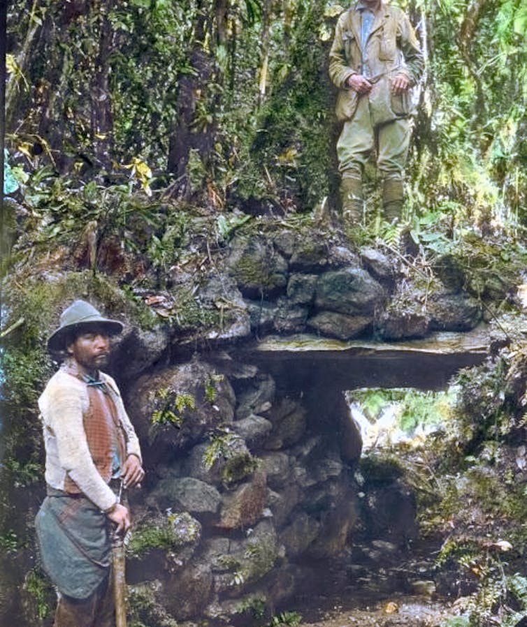 Un hombre vestido de explorador en lo alto de un puente mientras otro hombre mira a cámara desde el camino de debajo.